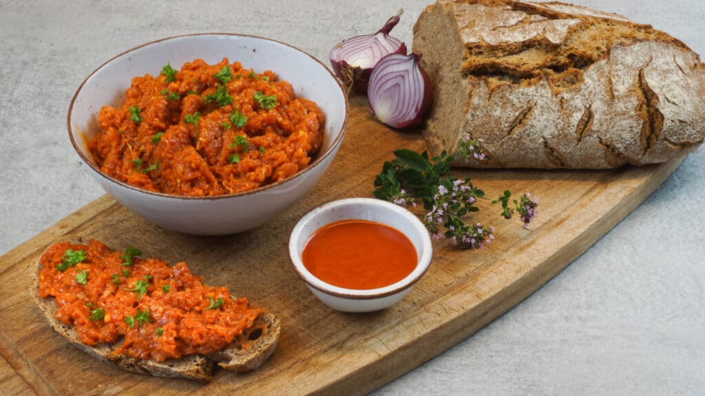 Metzgermett mit Würzsauce in einer Schüssel und auf einer Scheibe Brot + pure Würzsauce in kleiner Schüssel + ein angeschnittenes Laib Brot angerichtet auf einem Holzbrett