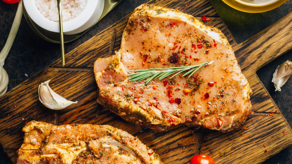 Mariniertes, rohes Fleisch mit Gewürzen auf einem Holzbrett, versehen mit einer Zwiebel und Tomaten